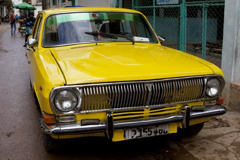 A bright yellow vintage car with a chrome grille parked beside a green fence