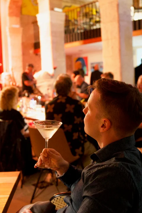 Me holding a cocktail glass in the warmly lit jazz club, with other guests behind me