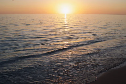 Gentle waves reaching a sandy shore beneath the setting sun and its reflection on the sea