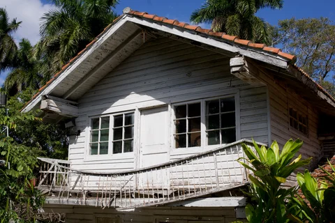 A white wooden building with a weathered balcony and red tiled roof among palms