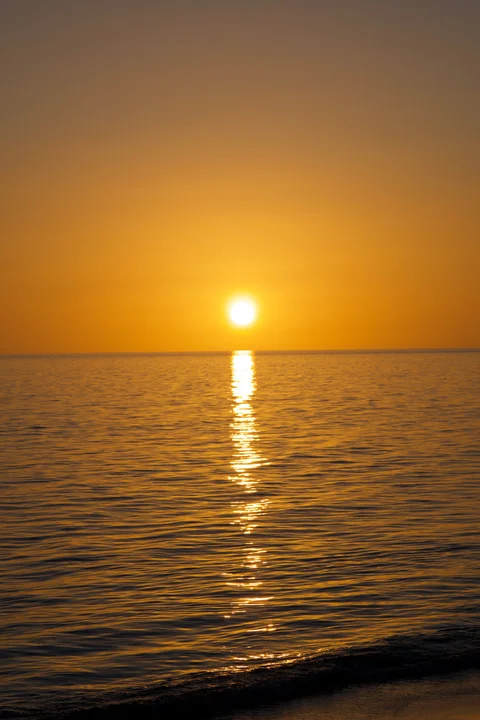 The setting sun and its bright golden reflection across the water, with a small wave reaching the shore