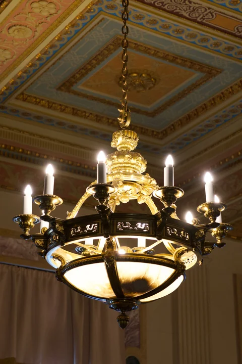 A glowing chandelier beneath a painted and gilded ceiling inside the Capitol