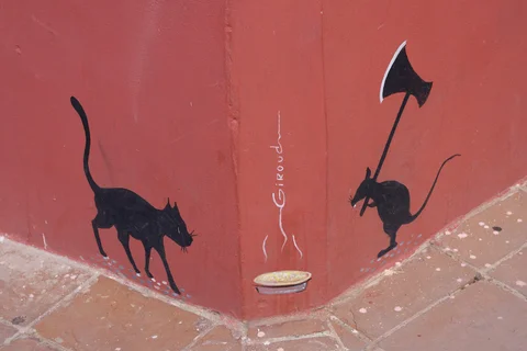 A black cat and a mouse carrying an axe painted around a red wall corner, with a bowl between them