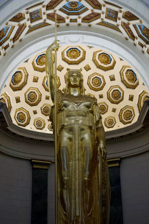 A gilded female statue holding a spear beneath an ornate ceiling inside the Capitol