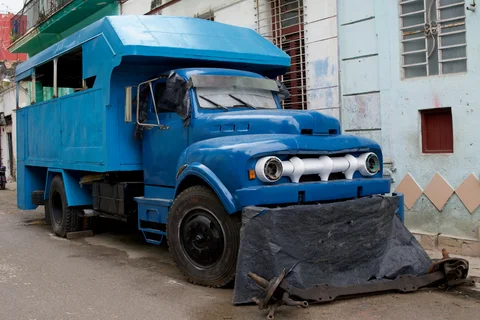 An old blue truck with a high passenger compartment and a tarpaulin in front of its bumper