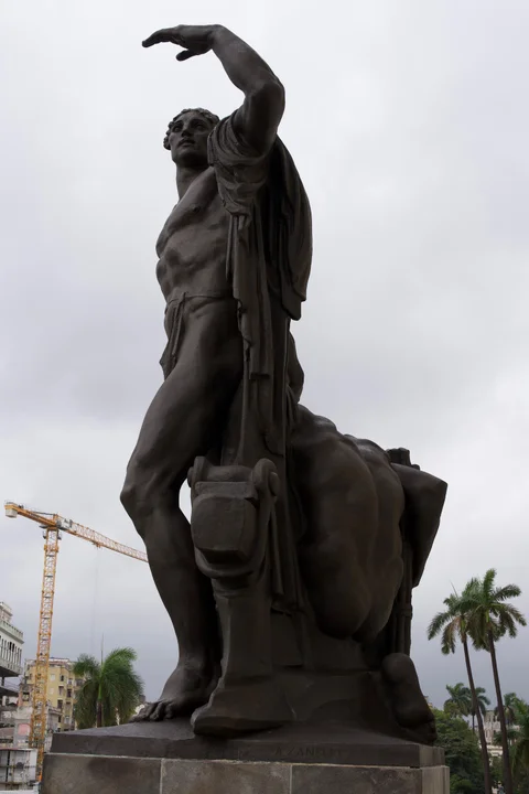 A large bronze male figure with one arm raised outside the Capitol, with palms and a crane behind it