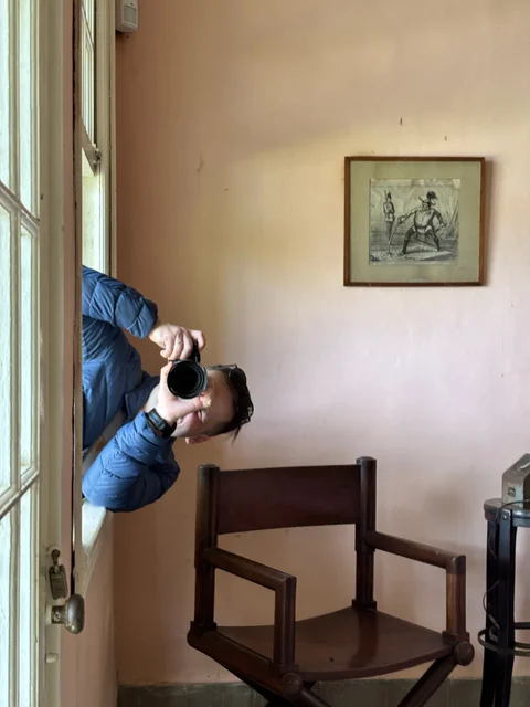 Me leaning through an open window with a camera, beside a wooden chair and framed print