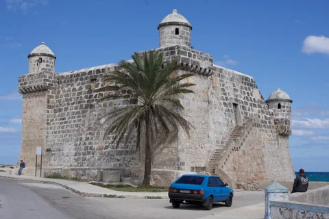 The stone walls and corner turrets of the Torreón de Cojímar, with a palm and a blue car in front