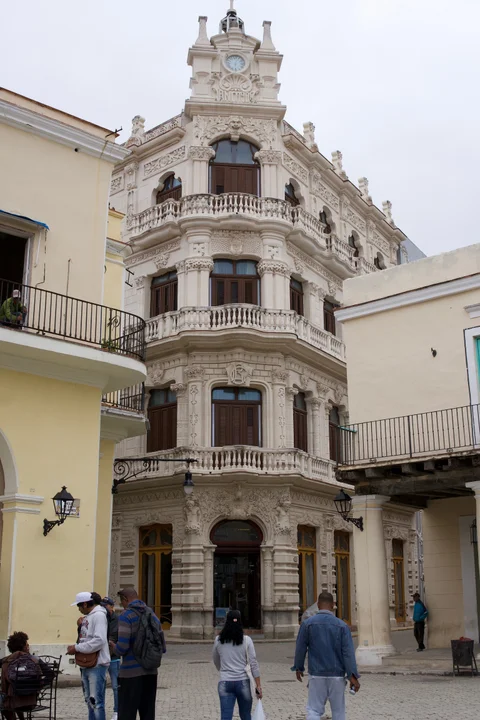 An ornate pale corner building with carved balconies and a clock above a Havana street