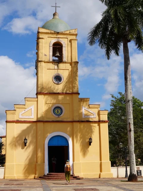 A yellow church with a blue doorway and a bell tower beside a tall palm in Viñales