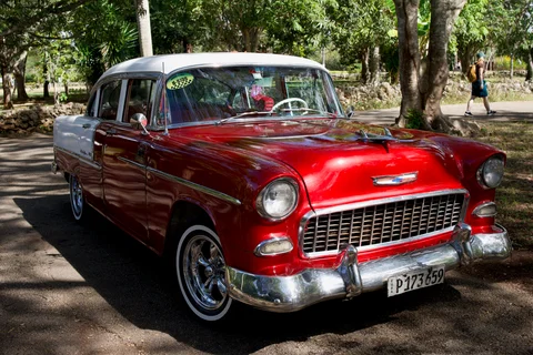A red and white vintage Chevrolet with polished chrome parked in the shade of trees