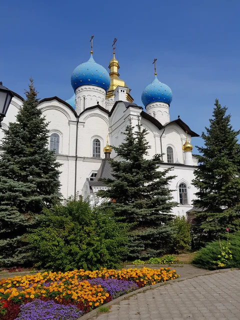 Blue domes of the Annunciation Cathedral above evergreens and a flower bed.