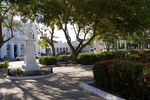 Paths, clipped hedges and a marble bust in Parque José Martí, with pastel buildings and an arch behind them