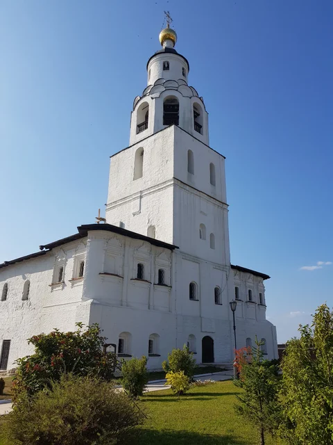 A white church and bell tower in Sviyazhsk, surrounded by grass and flowers.