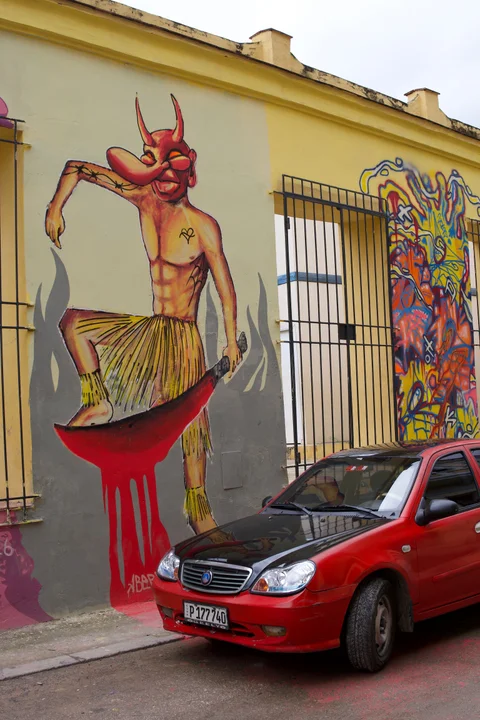 A red horned figure painted on a yellow wall above a parked red car