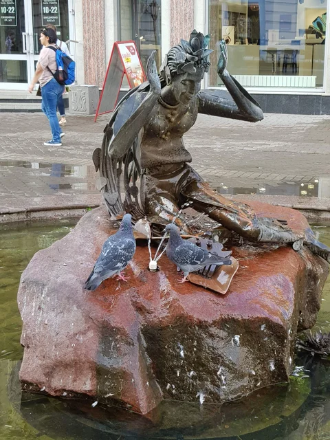 A bronze female figure sits on a red stone in a street fountain, with pigeons beside her.