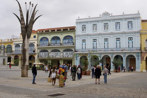 People crossing a Havana square lined with pastel facades, beside a tall metal plant sculpture