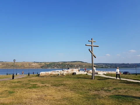 A wooden Orthodox cross stands on open grass above the water.