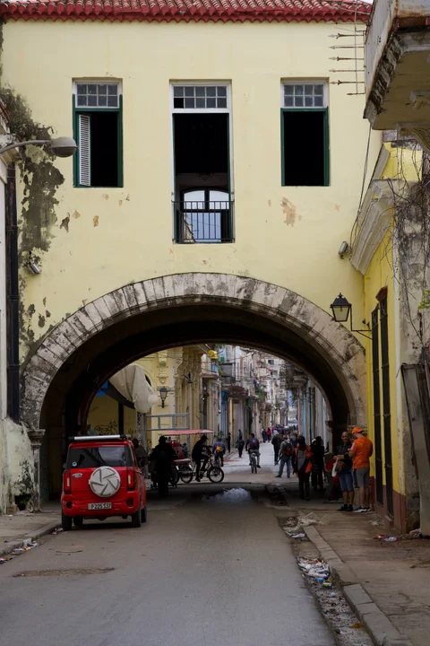 A stone arch beneath a yellow building spanning a street with people and a small red car