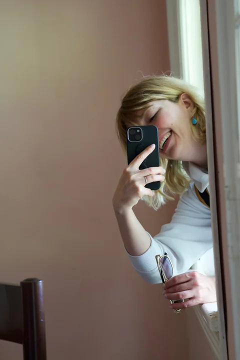 Lena smiling as she leans through an open window with her phone