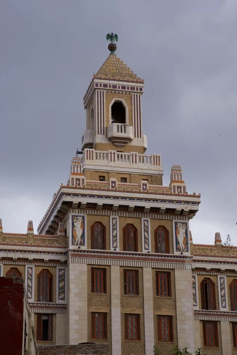 An ochre tower with white trim, topped by a bat figure on a globe, beneath grey clouds