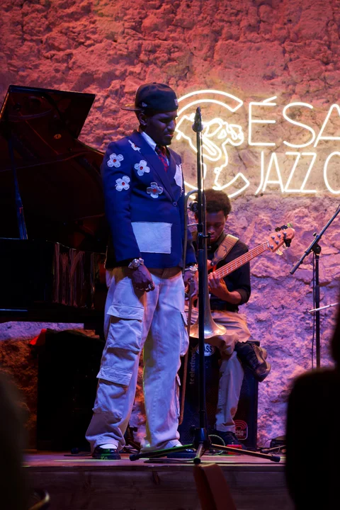 A performer at a microphone in front of the illuminated club sign, with a bass guitarist behind him