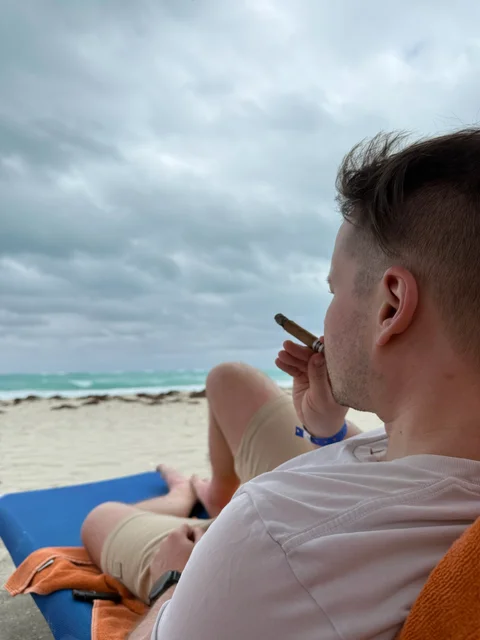 Me reclining on a blue beach lounger with a cigar beside the sea under a cloudy sky