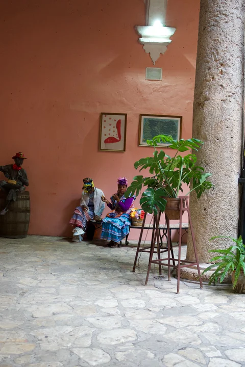 Two women in colourful dresses seated beside plants and a stone column in the Havana Club courtyard