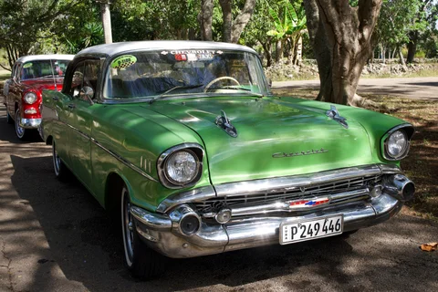 A green vintage Chevrolet with a white roof and chrome bumper, with a red car behind it