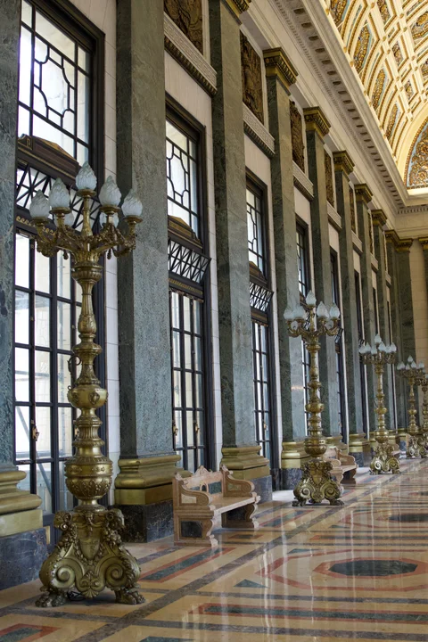Ornate floor lamps and stone benches beside tall windows in the Capitol’s marble hall