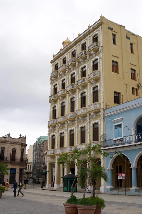 A tall yellow building with white balconies beside a blue arcade on a Havana square