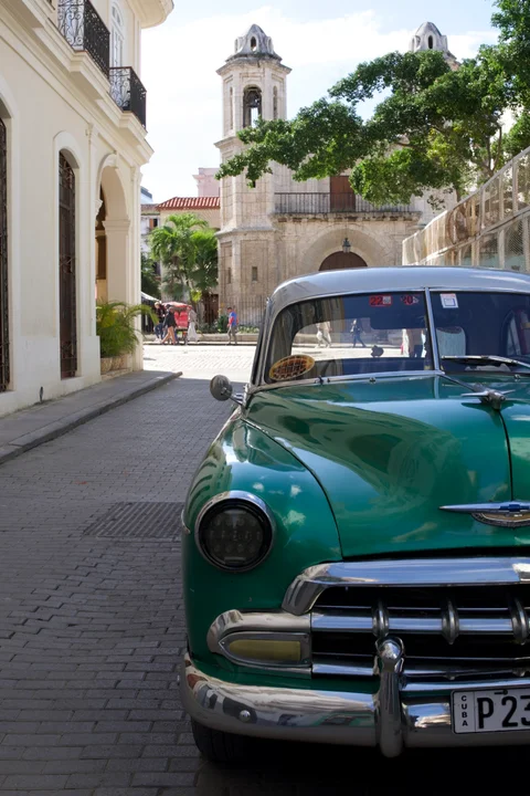 The front of a green vintage car beside a cobbled street, with a church tower beyond