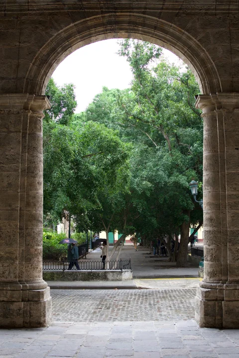 A stone arch framing leafy trees and pedestrians with umbrellas in Havana