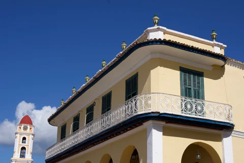 The yellow facade and white balcony of Palacio Brunet in Trinidad, with the San Francisco bell tower behind it