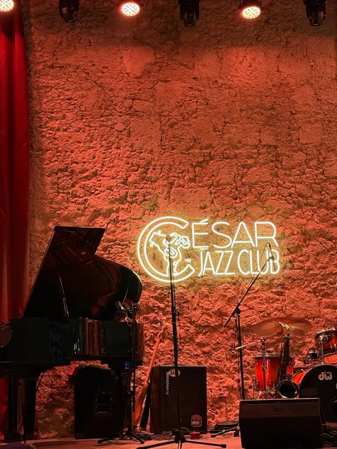 A piano and drum kit on the empty stage beneath the glowing César Jazz Club sign